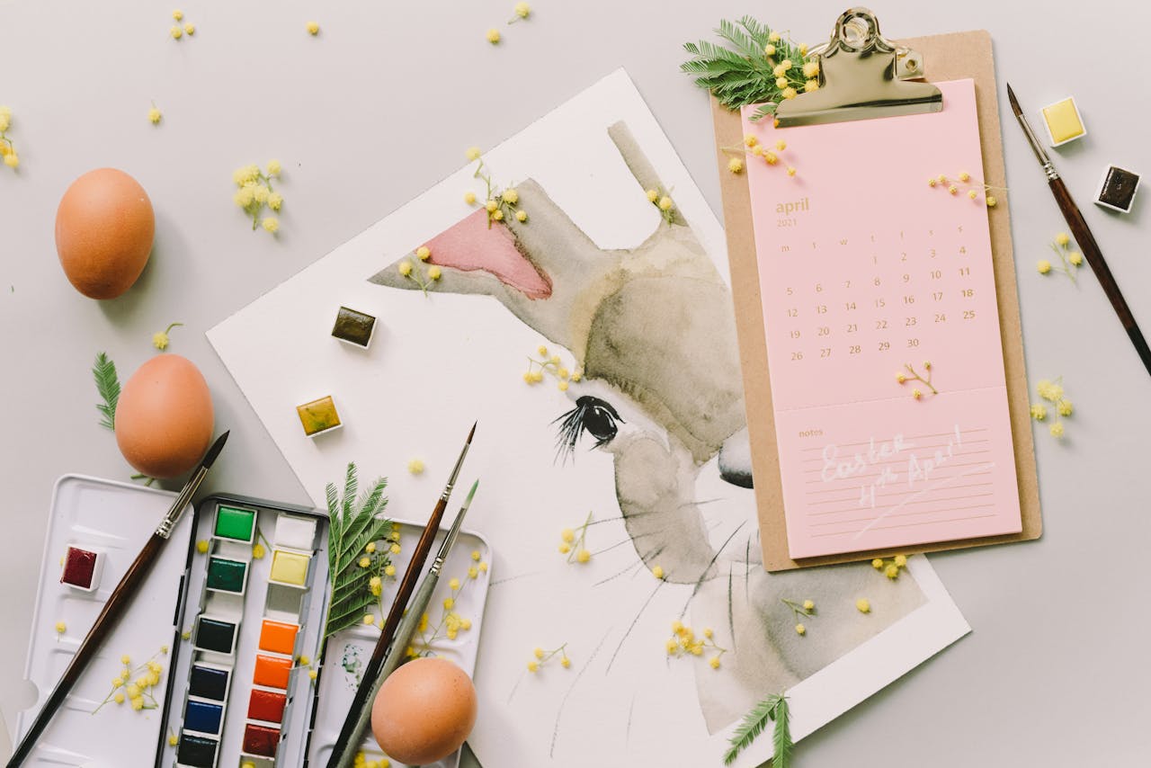 team-01 Flat lay of Easter bunny watercolor art, April calendar, eggs, and brushes for a creative spring celebration.